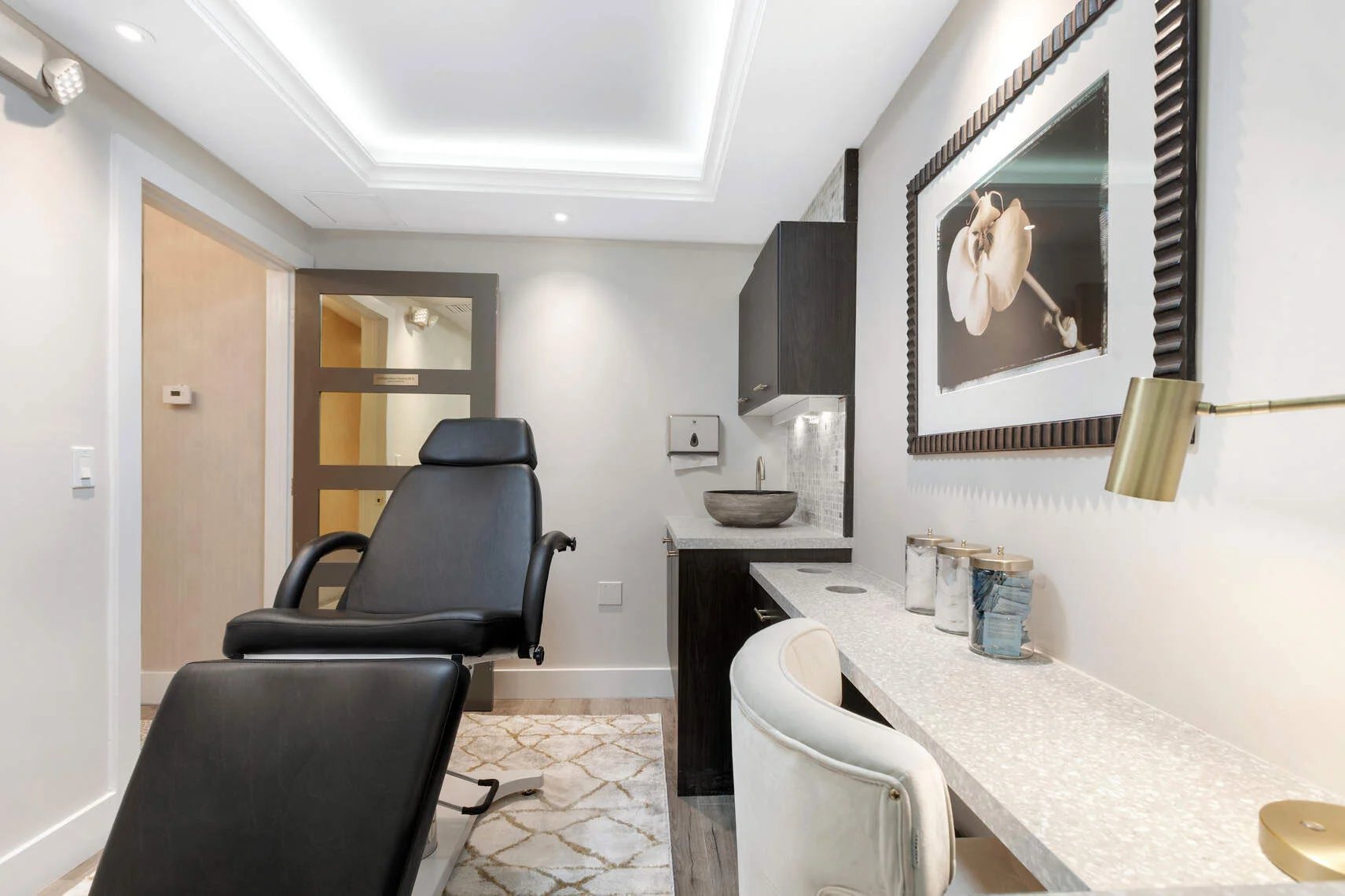Modern medical or spa treatment room with a black reclining chair, white desk chair, countertop with a stone sink, and framed orchid artwork on the wall.