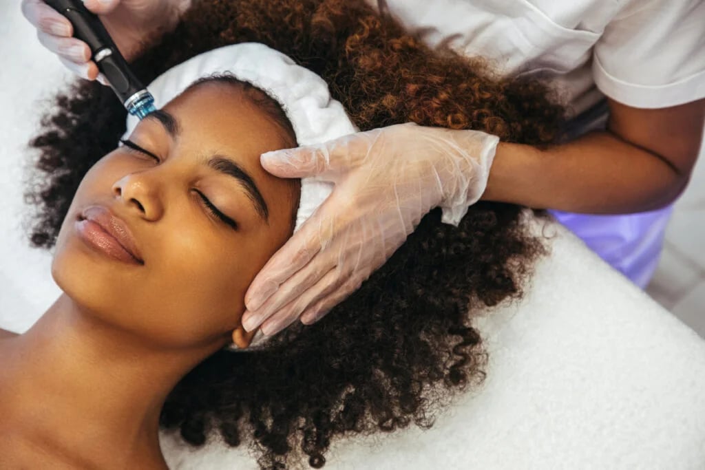 A woman with curly hair receiving a facial treatment while lying on a white towel with eyes closed.