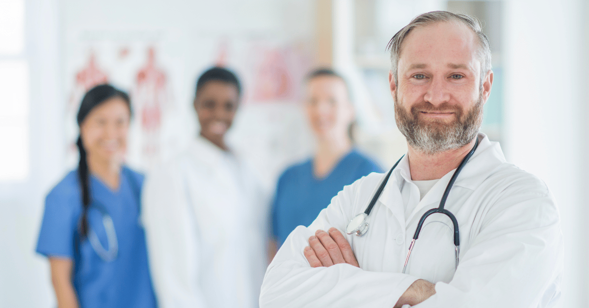 Smiling male doctor with beard and stethoscope standing with folded arms in front of three diverse medical professionals.