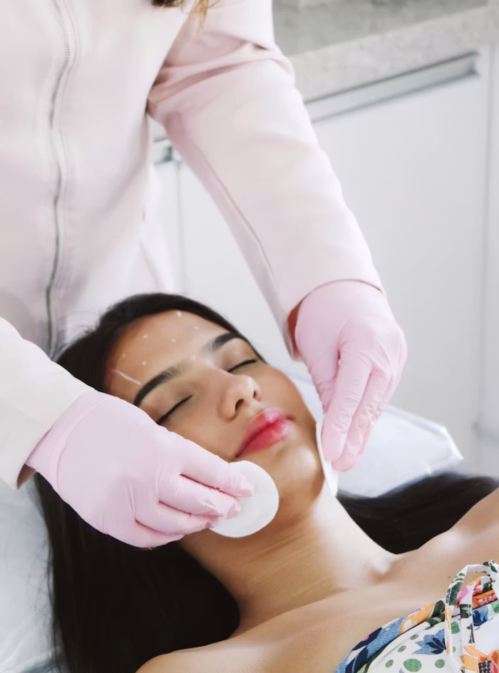A woman with closed eyes receiving a facial treatment as a person wearing pink gloves cleans her cheek with a cotton pad.