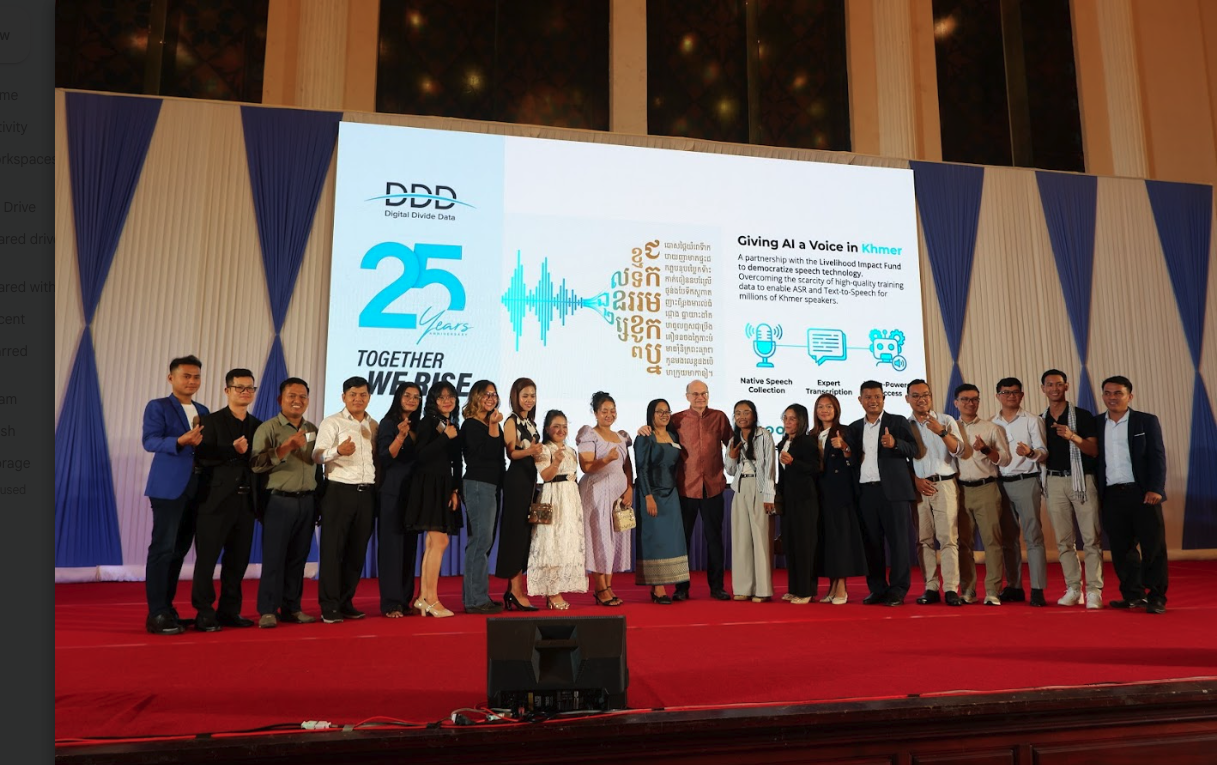 Group of people standing on a stage in front of a large display celebrating 25 years of Digital Divide Data and promoting AI voice technology for Khmer language.