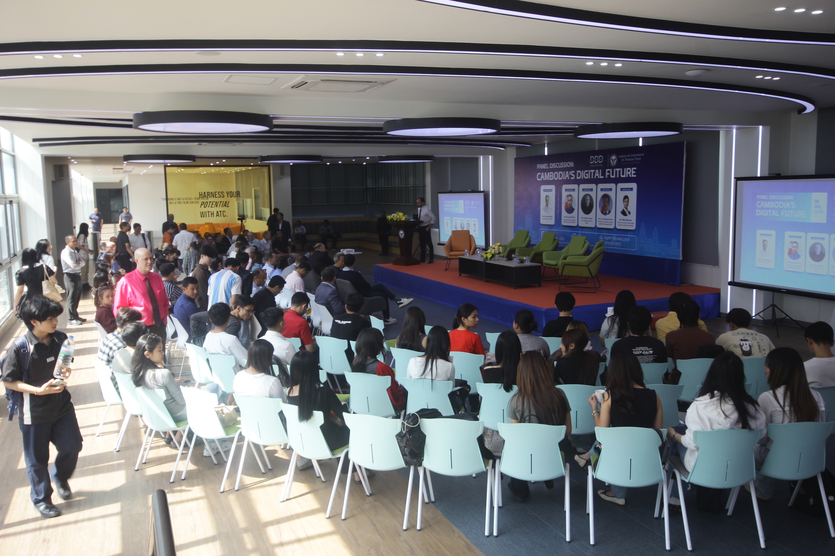 Audience at a panel discussion event on Cambodia's digital future, seated facing a stage with a speaker at a podium and large screens displaying panel information.