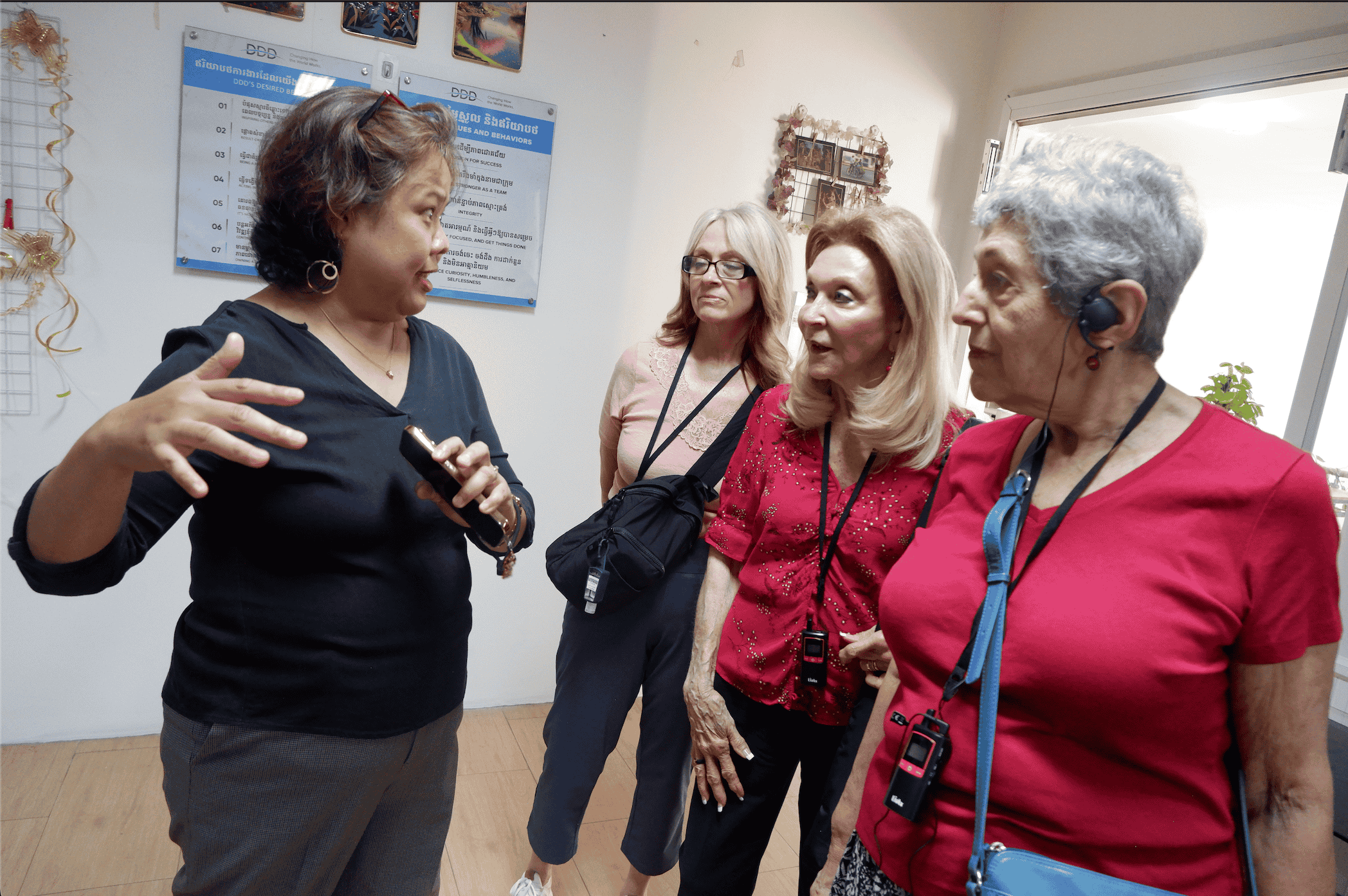 A woman in black explaining something with hand gestures to three attentive older women wearing visitor badges in an indoor setting.