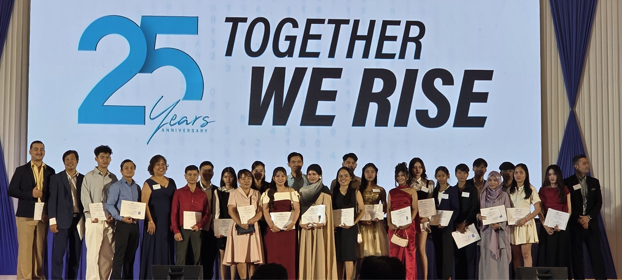 Group of people standing on stage holding certificates in front of a screen displaying '25 Years Anniversary' and 'Together We Rise' text.