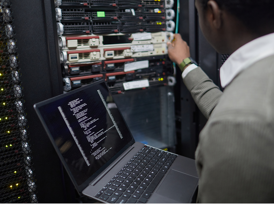 Technician in a server room working on network equipment with a laptop displaying code.