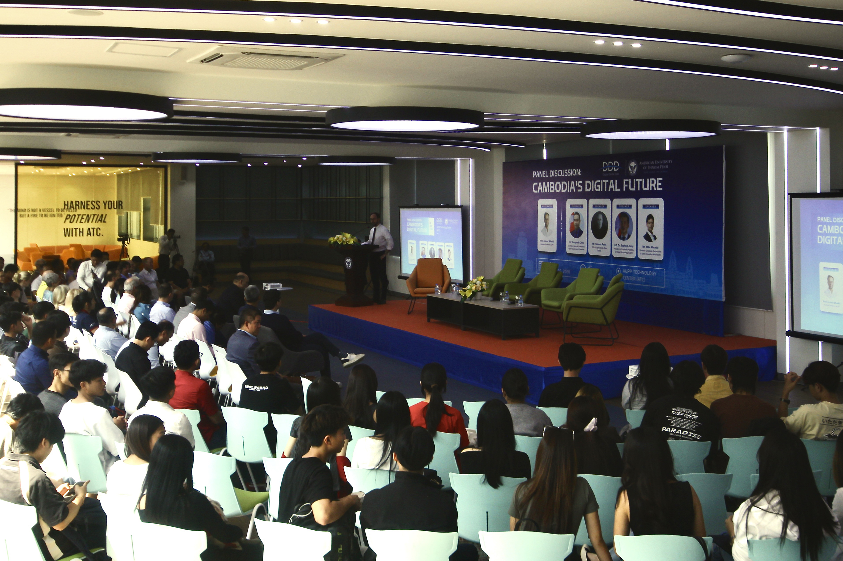 Audience at a panel discussion event on Cambodia's digital future, seated facing a stage with a speaker at a podium and large screens displaying panel information.