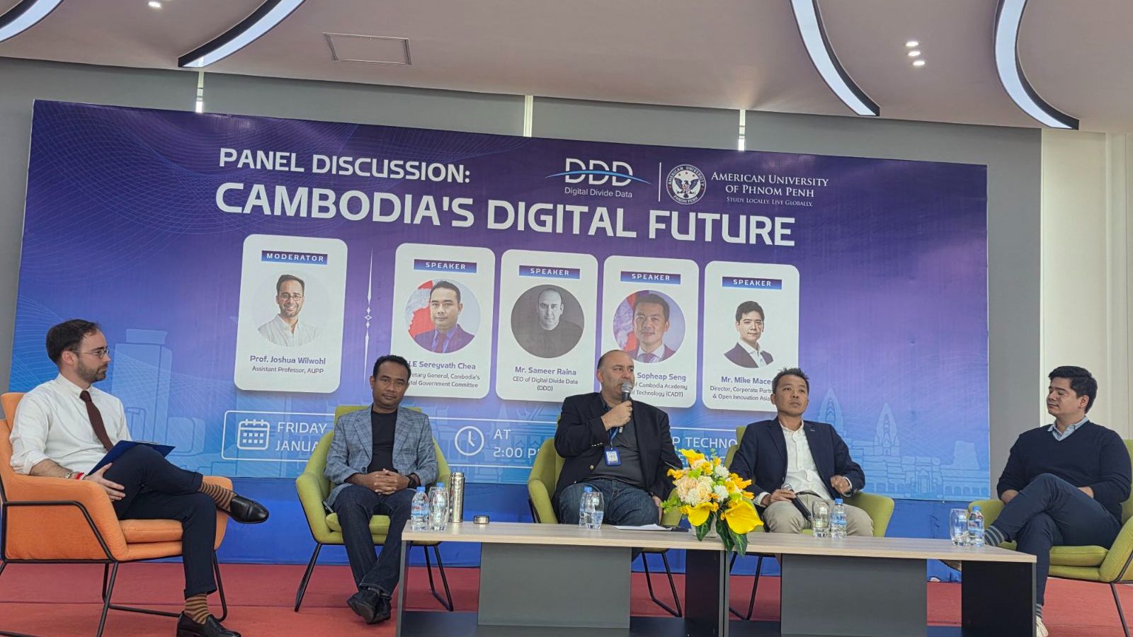 Audience at a panel discussion event on Cambodia's digital future, seated facing a stage with a speaker at a podium and large screens displaying panel information.