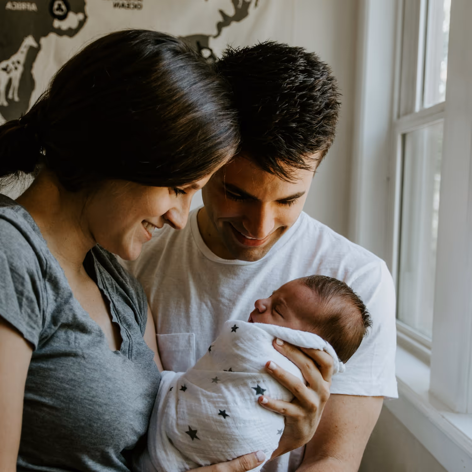 Casal em sua casa, lado a lado, a olhar para o seu filho bebé, que está nos braços da mãe.