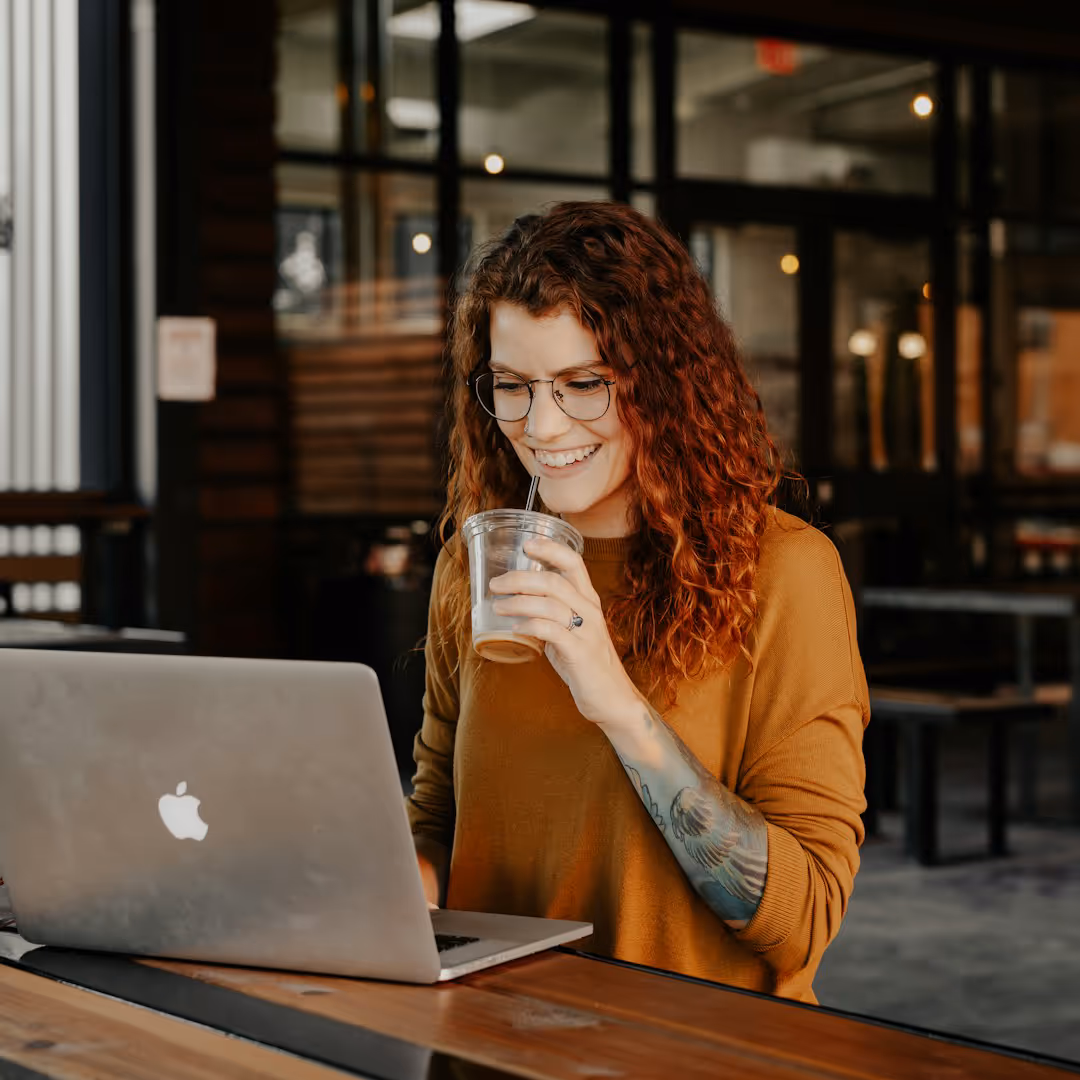 Jovem a utilizar computador, numa cafetaria, com uma bebida na mão, a sorrir.