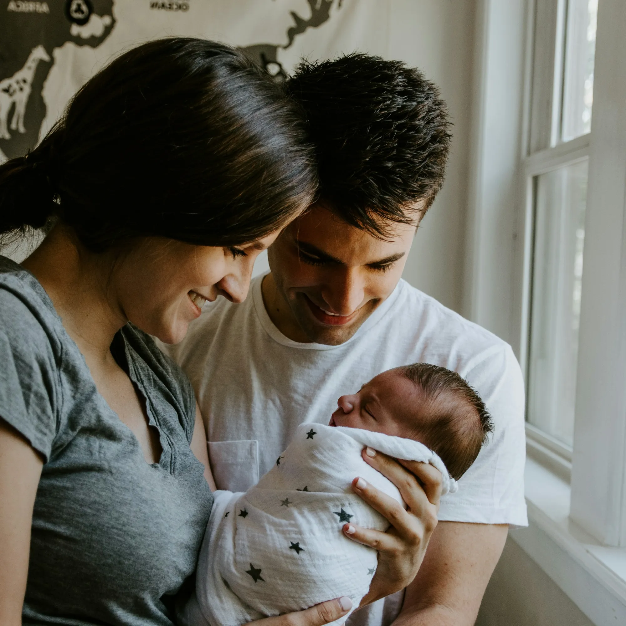 Casal em sua casa, lado a lado, a olhar para o seu filho bebé, que está nos braços da mãe.