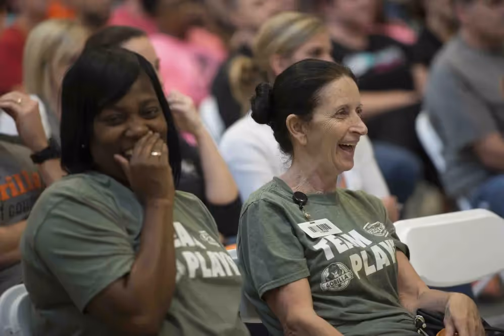 Two women laughing and sitting in an audience at an event, wearing green shirts with a logo and text.