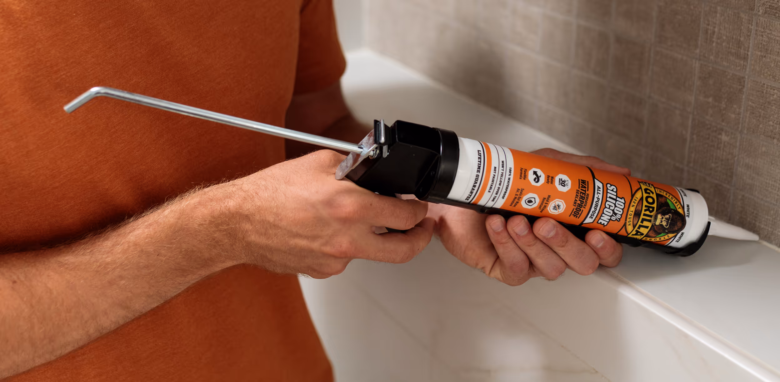 Person holding a tube of Gorilla 100% Silicone Sealant with an attached caulking gun near a tiled surface.