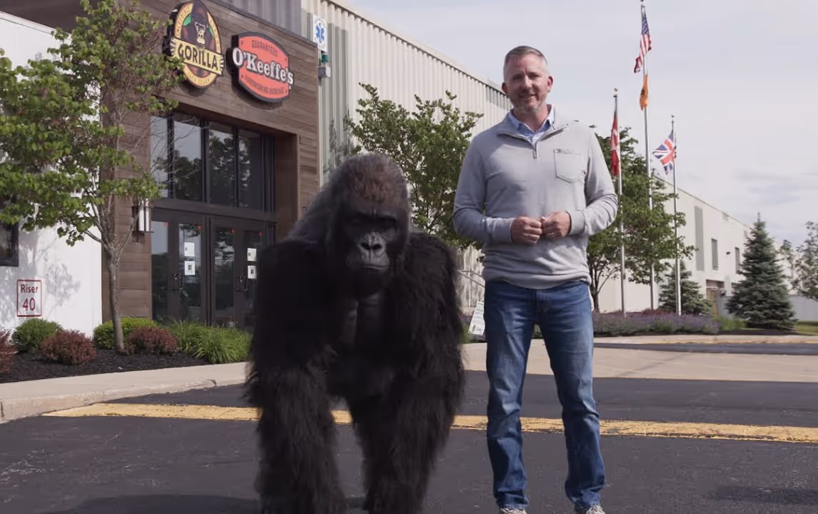 Man standing outdoors next to a large realistic gorilla statue in front of a building with O'Keeffe's and Gorilla logos.