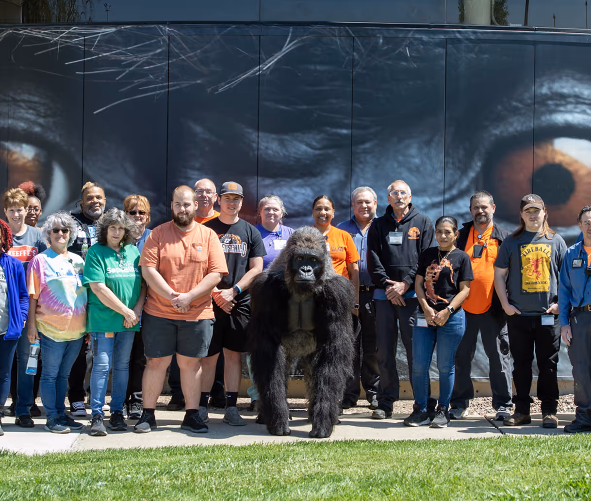 Group of diverse people posing outdoors with a realistic gorilla standing in front of a large mural of gorilla eyes.