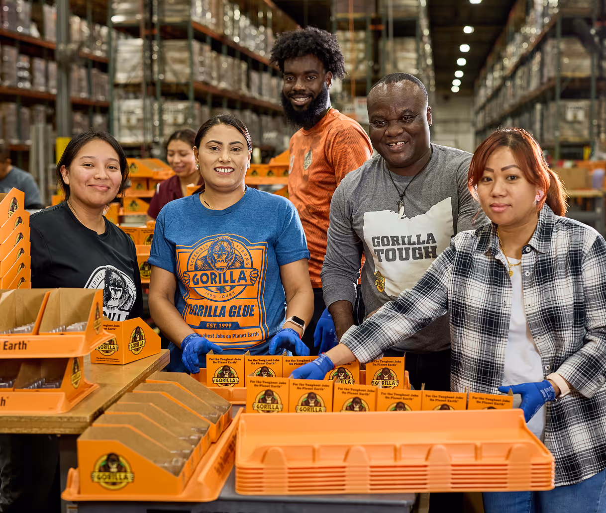 Five diverse workers smiling and packing orange Gorilla Glue boxes in a warehouse.
