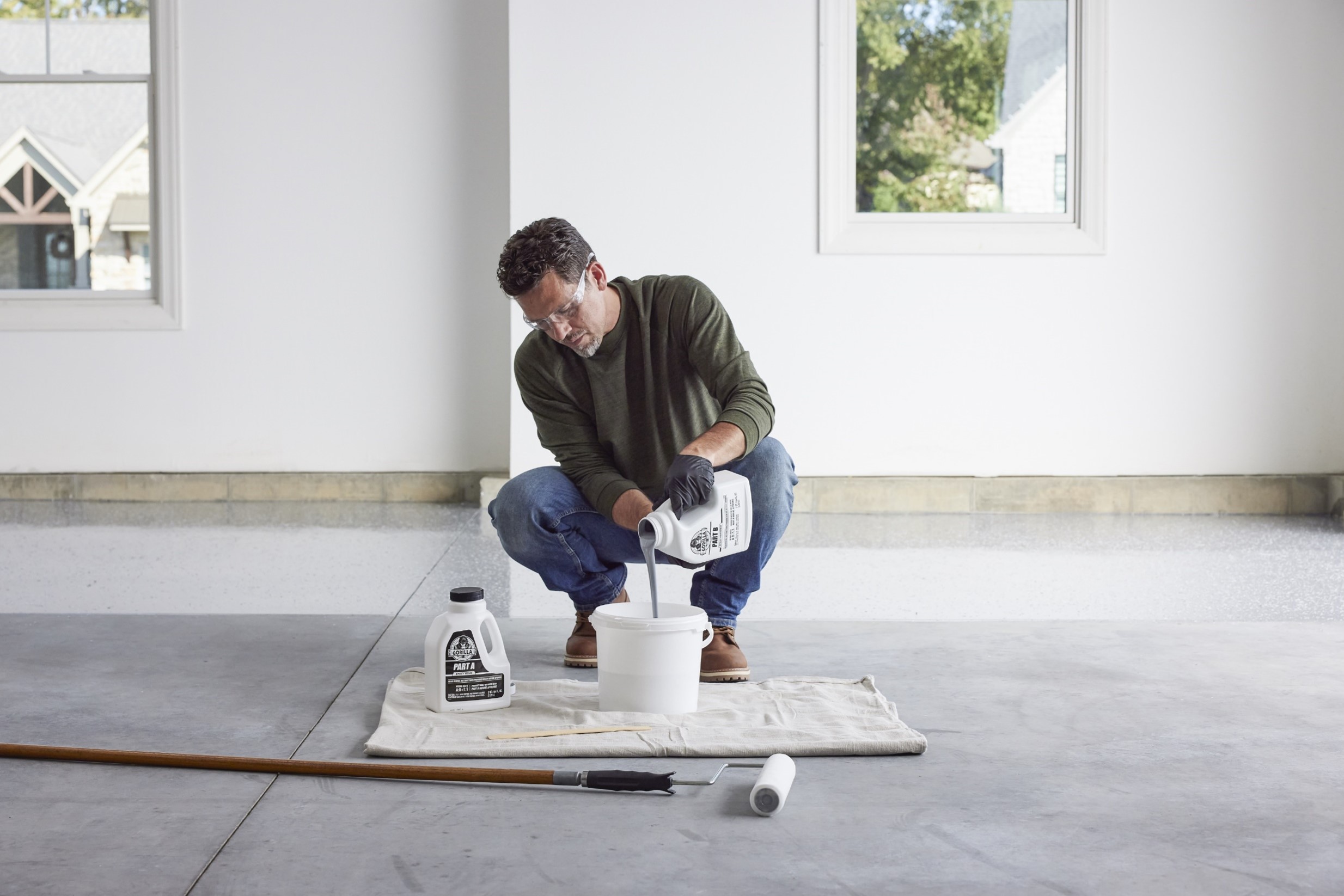 Man wearing safety glasses and gloves pouring liquid from a container labeled 'Part B' into a white bucket on a cloth in a bright room with two windows.