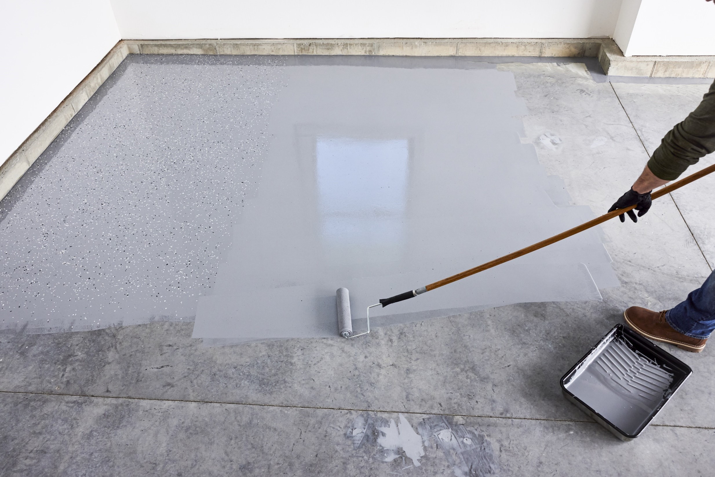 Person applying gray epoxy coating with paint roller on concrete garage floor.