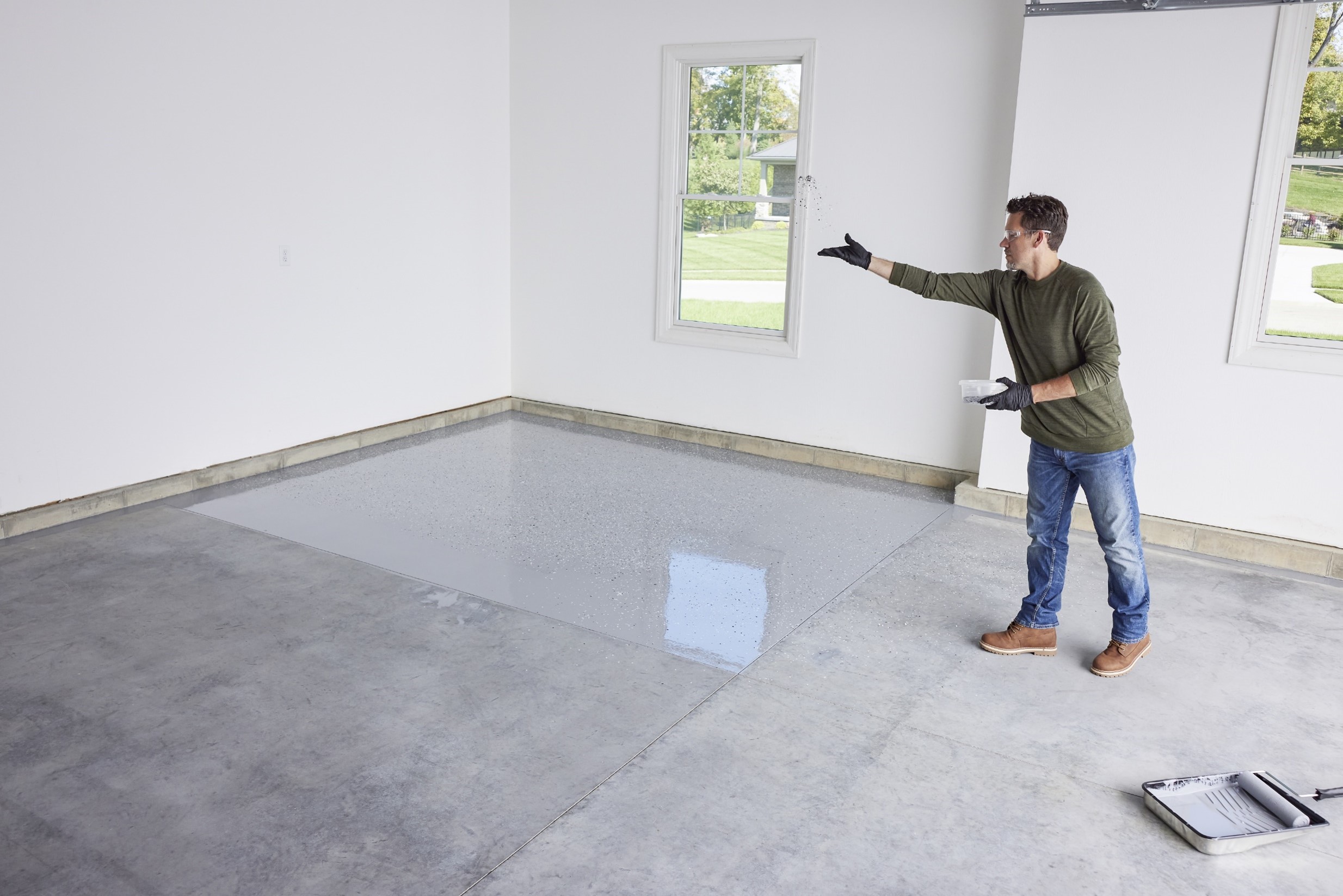 Man wearing gloves and safety glasses tossing decorative flakes onto a freshly coated epoxy garage floor.