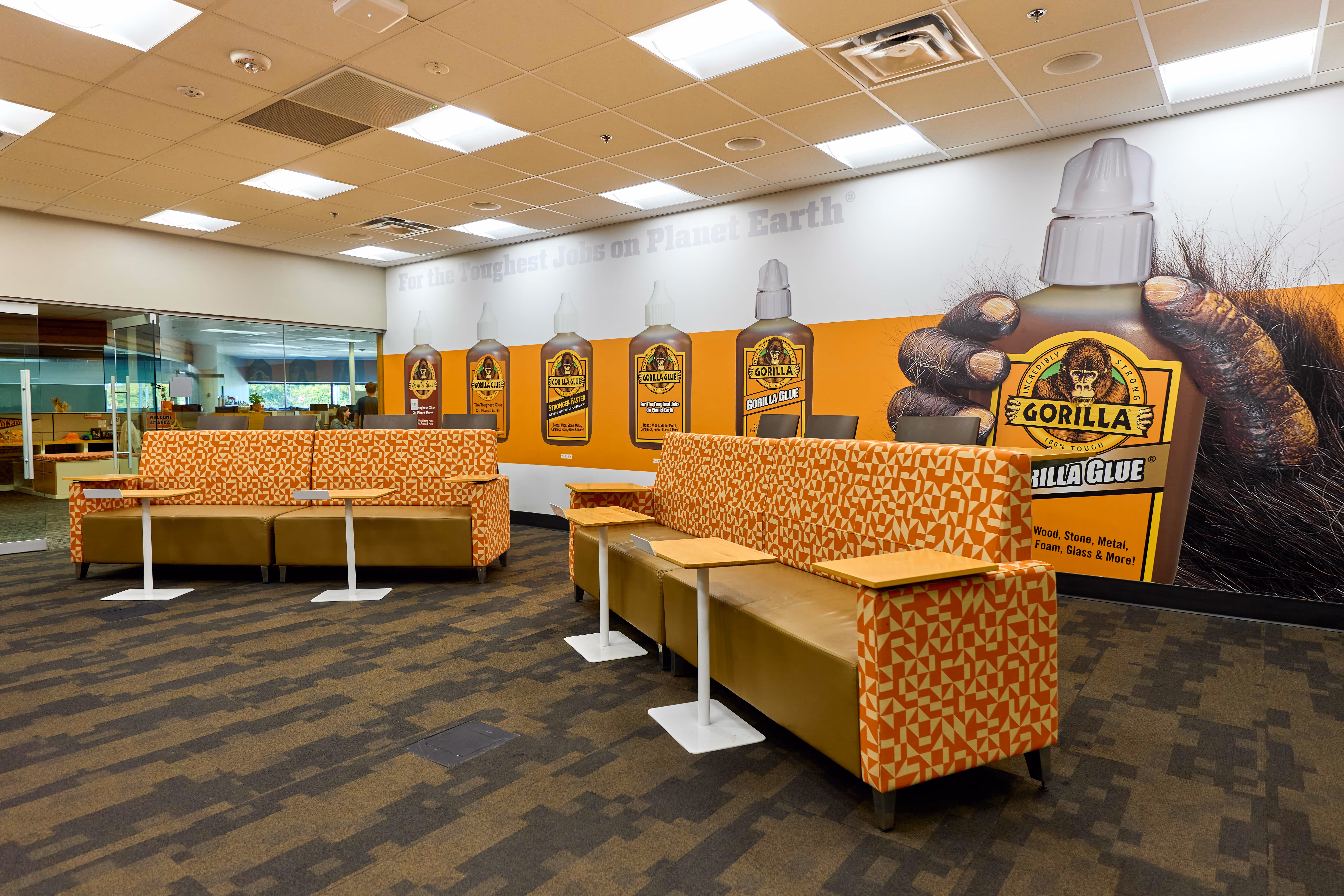 Office lounge area with patterned orange and beige sofas, small wooden tables, and a large wall mural featuring various Gorilla Glue bottles and a gorilla hand holding a glue bottle.