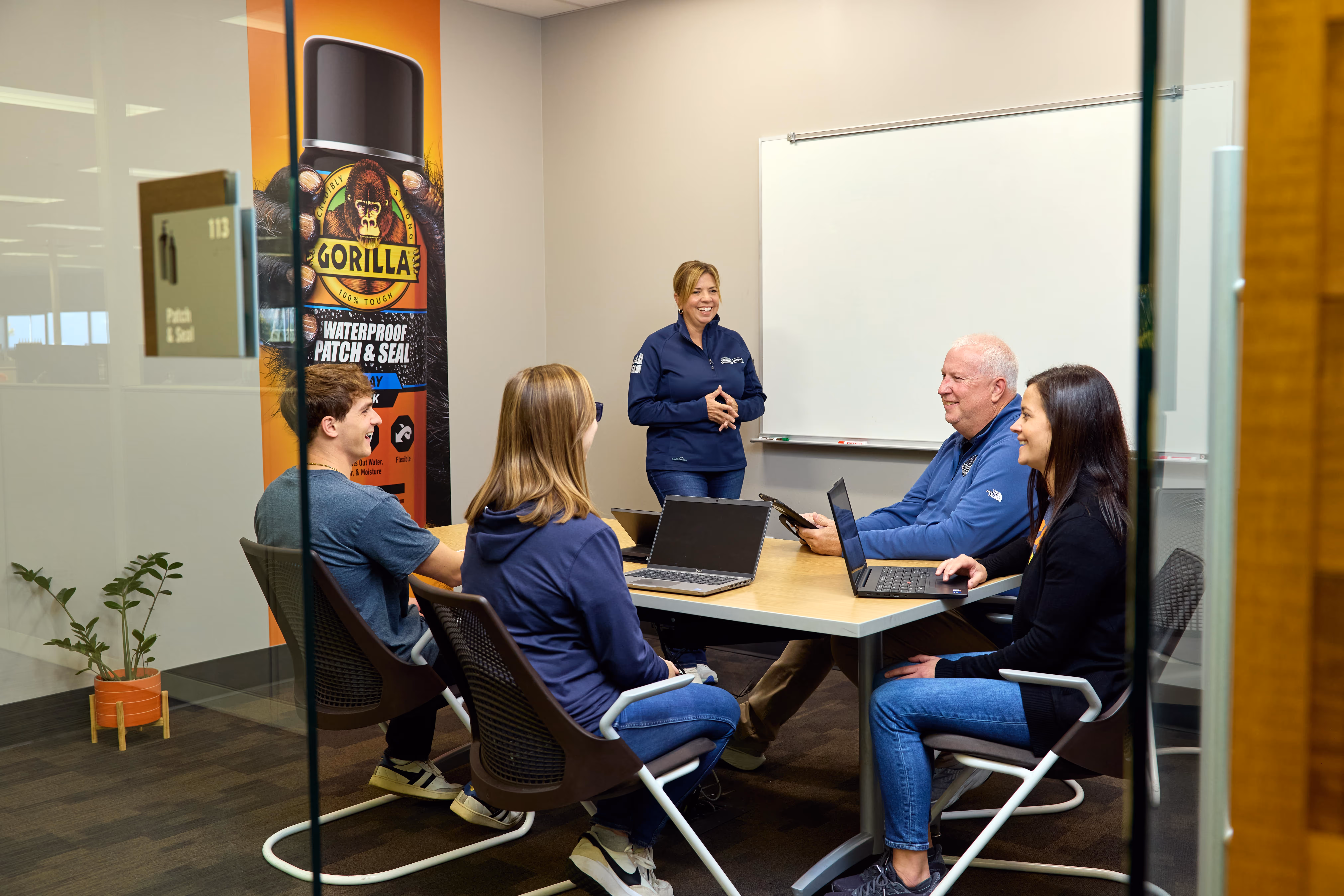 Five people in a conference room having a meeting with laptops and tablets, smiling and talking near a whiteboard and a Gorilla Glue advertisement.