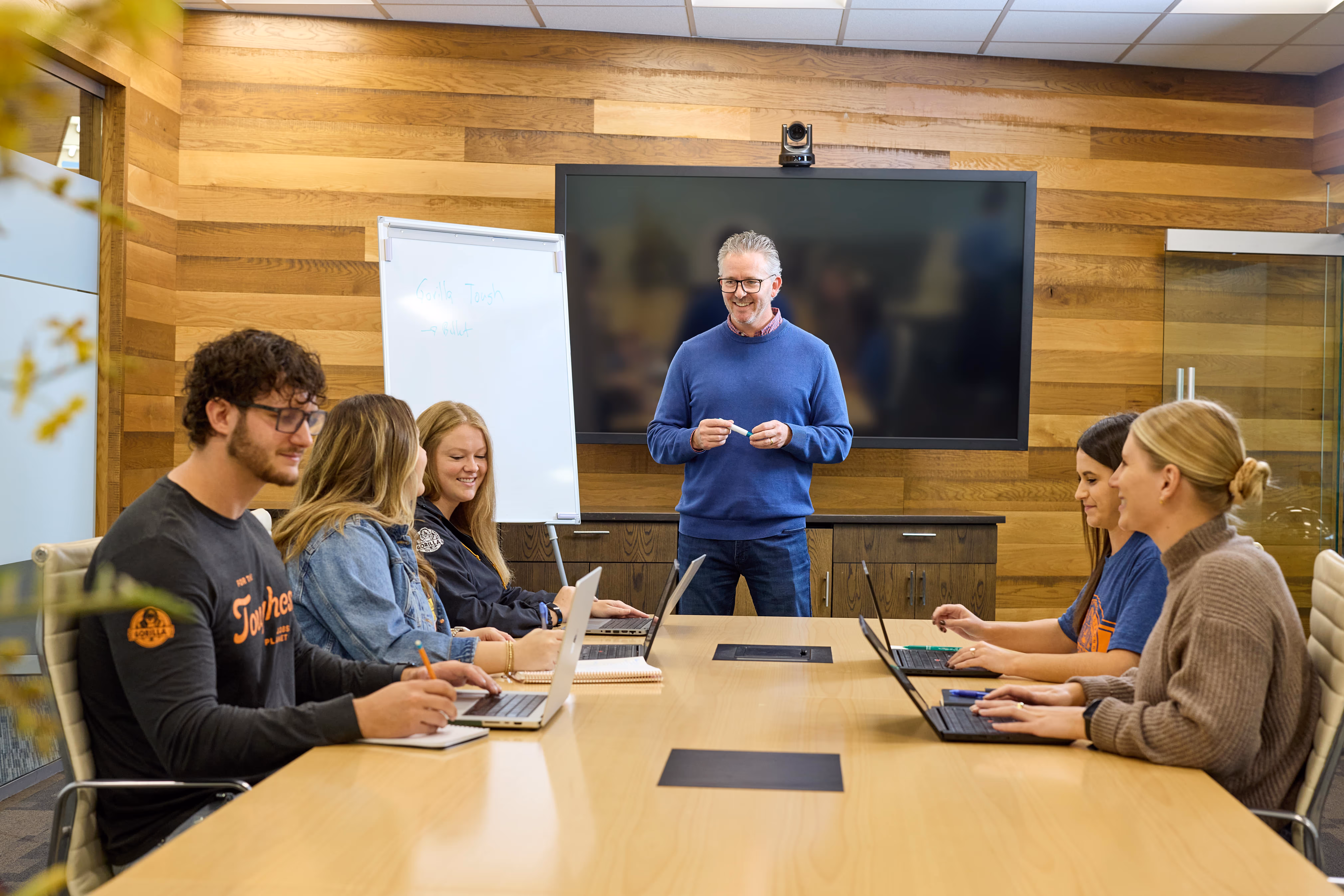 Man standing in a wood-paneled conference room leading a meeting with five people seated at a long table, using laptops and notebooks.
