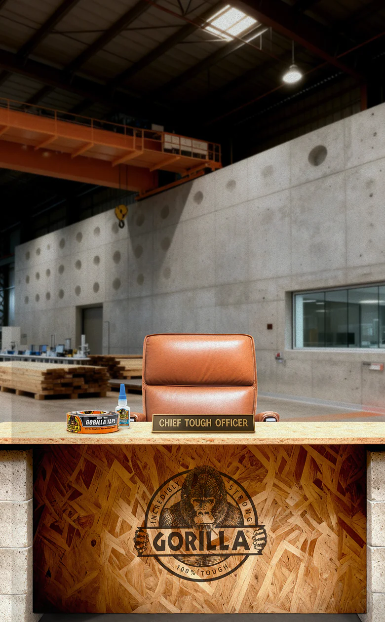 Empty brown leather office chair behind a wooden desk with a nameplate reading 'CHIEF TOUGH OFFICER' and Gorilla tape and glue on the desk; industrial warehouse background.