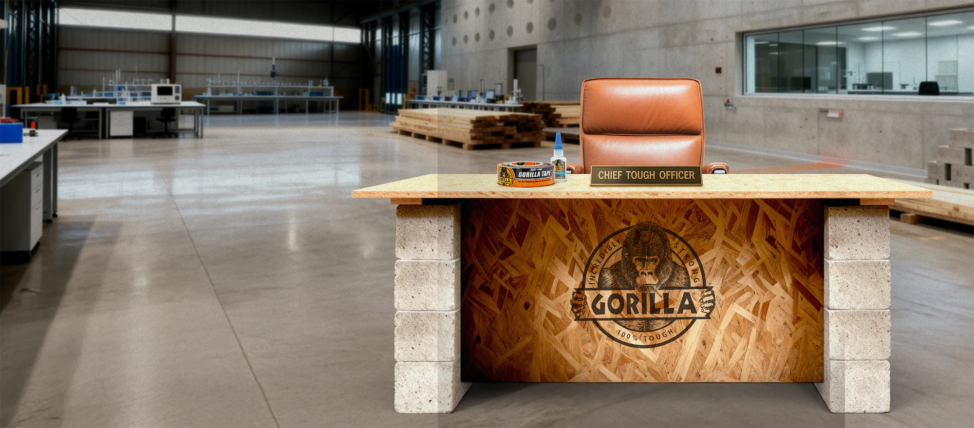 Office desk with cinder block legs and wood panel displaying Gorilla logo, topped with a leather chair, Gorilla tape, glue bottle, and a nameplate reading 'CHIEF TOUGH OFFICER' in a spacious industrial workshop.