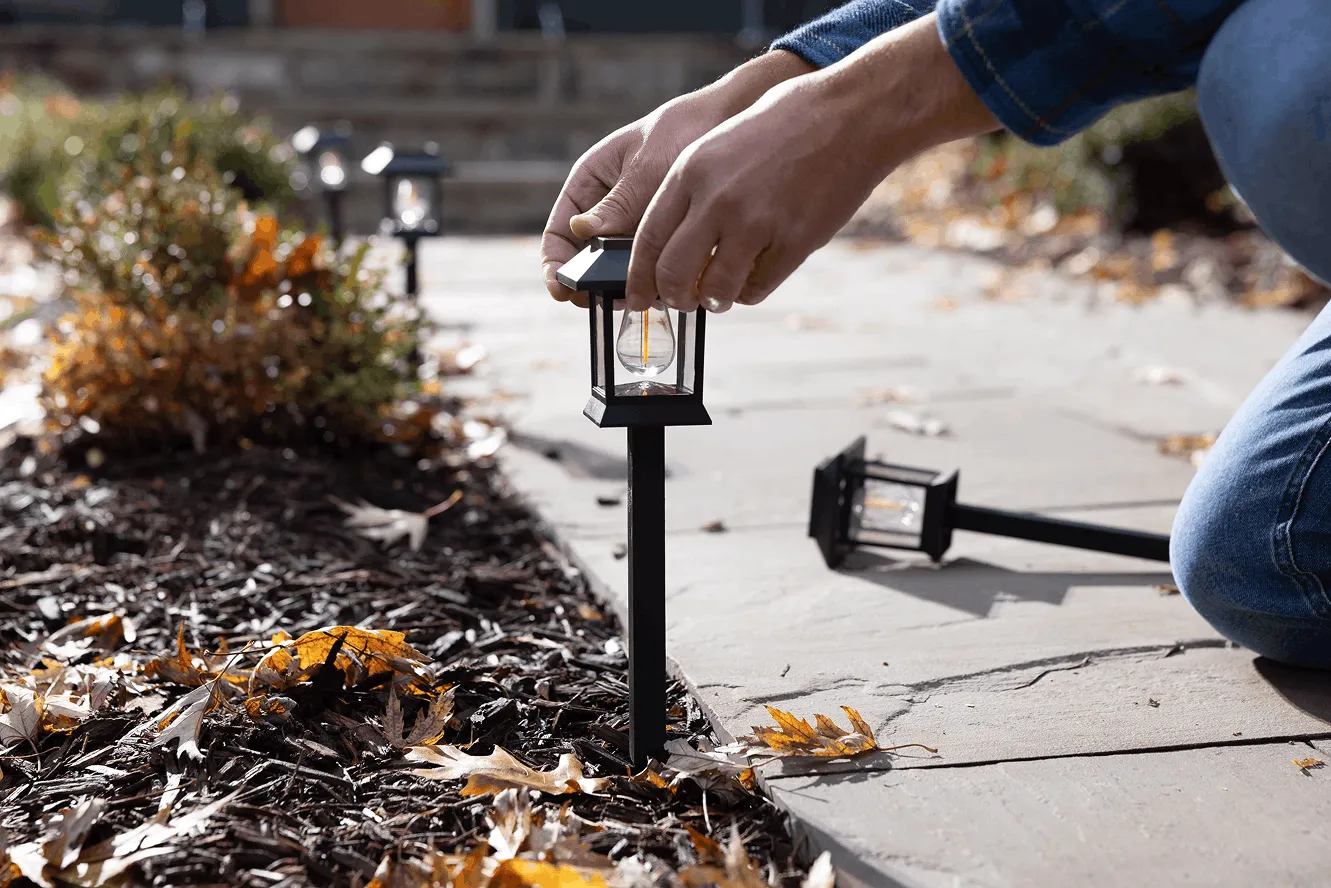 Person installing a black solar garden light along a stone pathway with autumn leaves on the ground.
