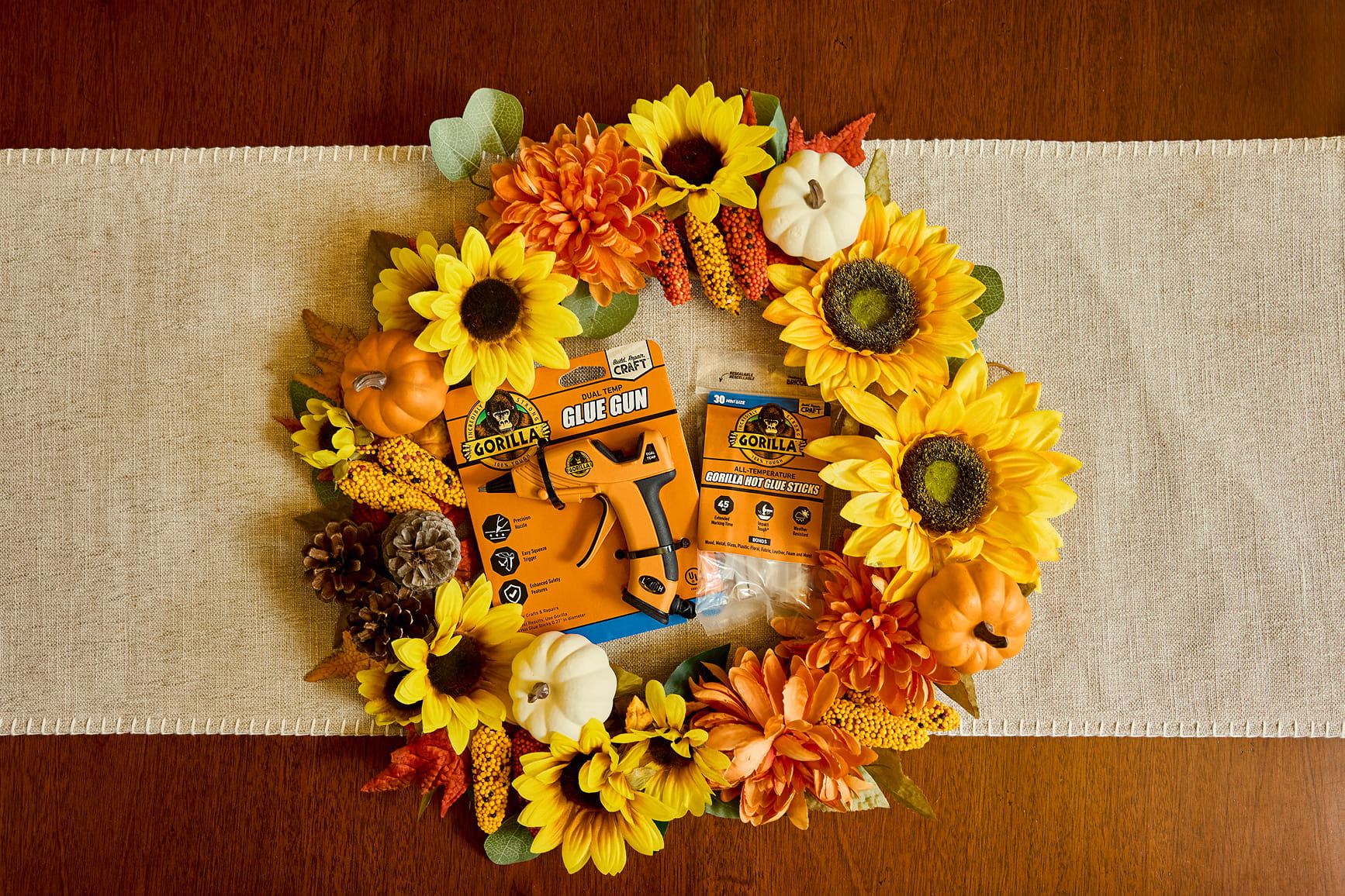 Autumn wreath with sunflowers, pumpkins, pine cones, and orange flowers surrounding Gorilla brand glue gun and hot glue sticks on a beige runner.