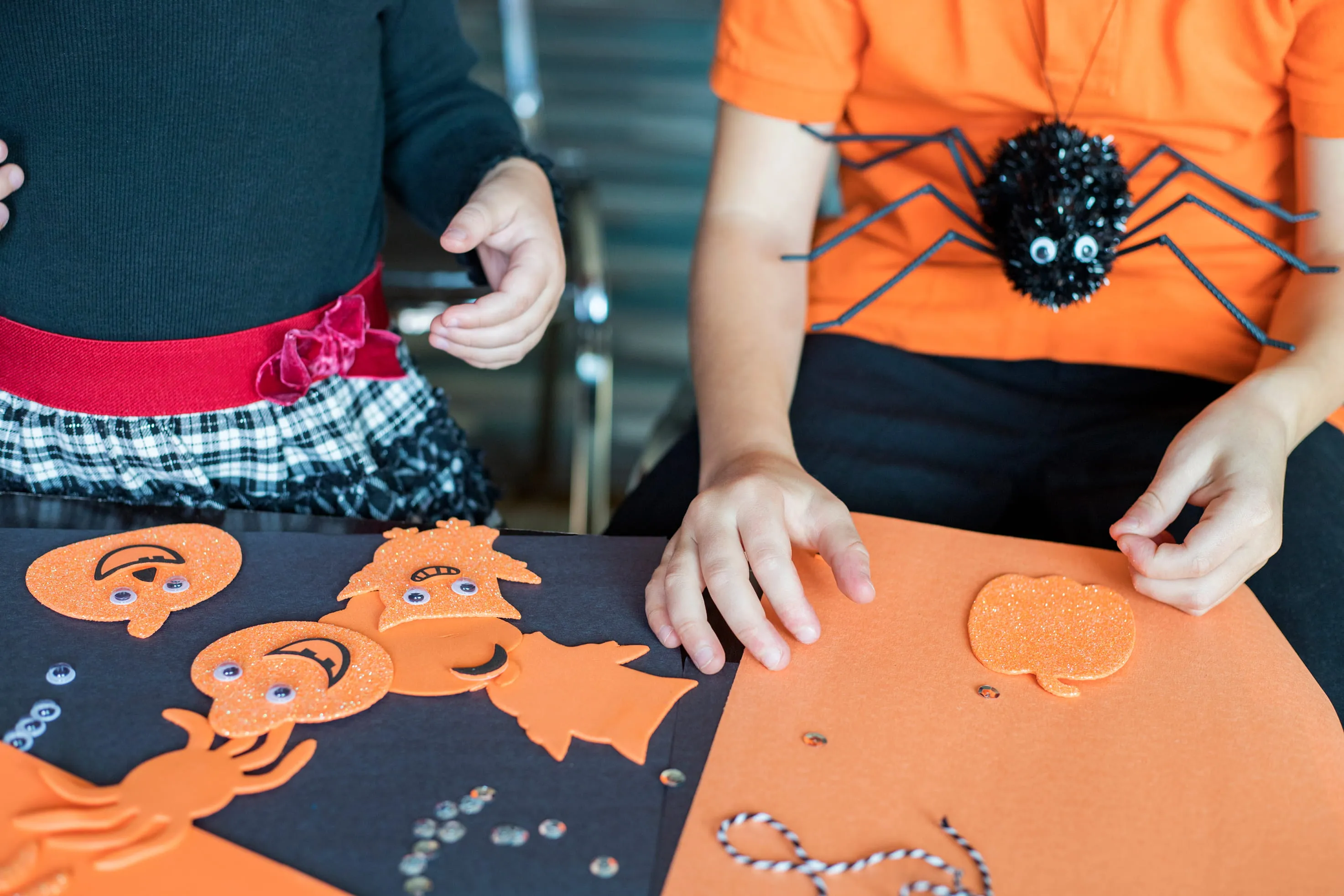 Two children crafting with orange Halloween-themed foam cutouts, one wearing an orange shirt with a large black spider decoration.