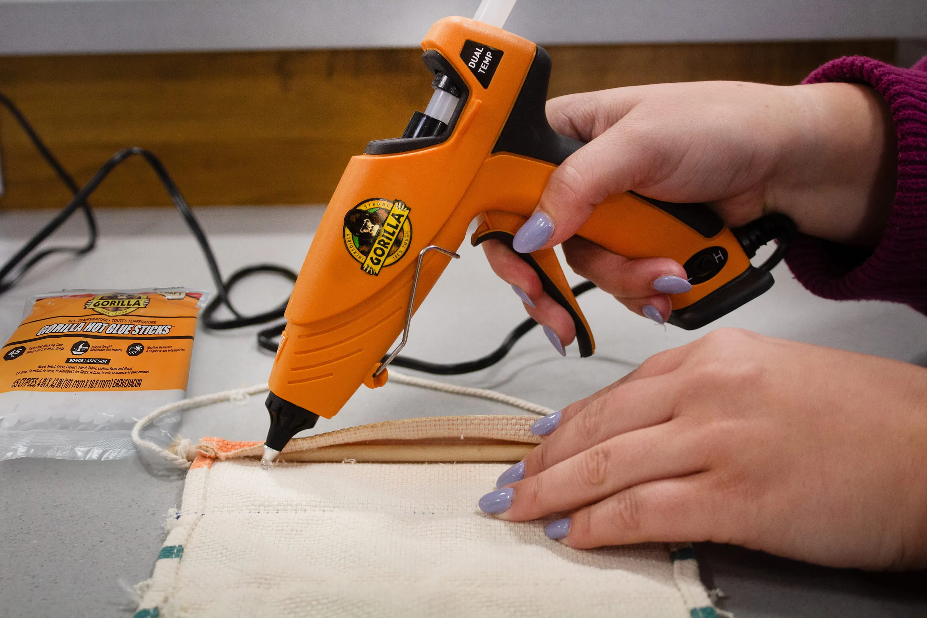 Person using an orange Gorilla hot glue gun to apply glue on a piece of fabric on a table.