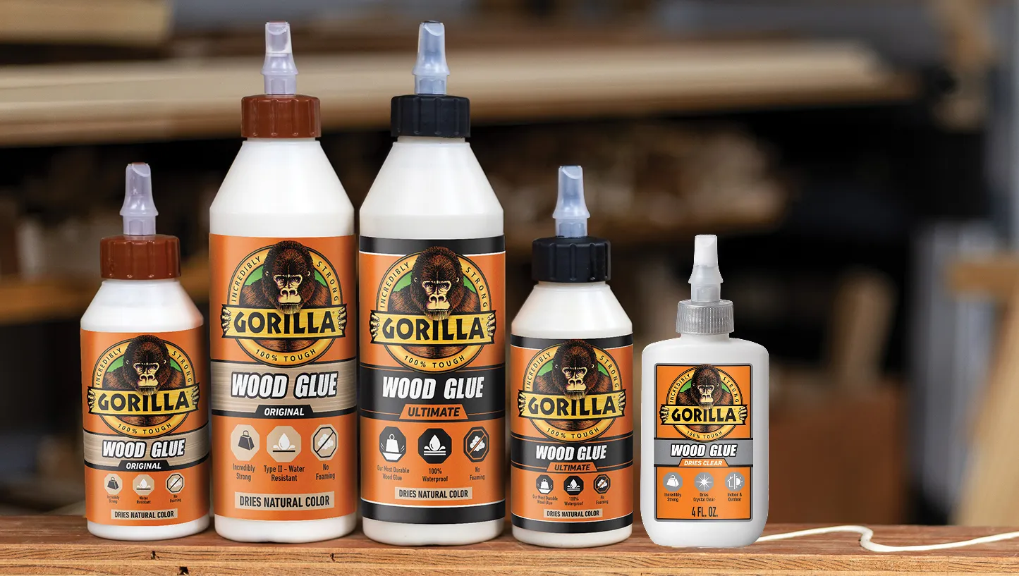 Five bottles of Gorilla wood glue in varying sizes and types, arranged on a wooden surface with a blurred woodworking background.