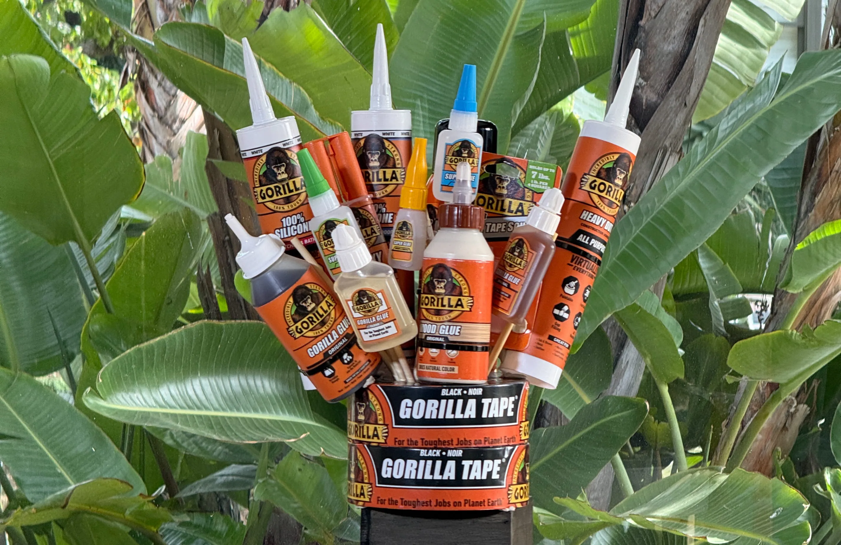 Variety of Gorilla brand adhesive products arranged in a container with tropical green leaves in the background.