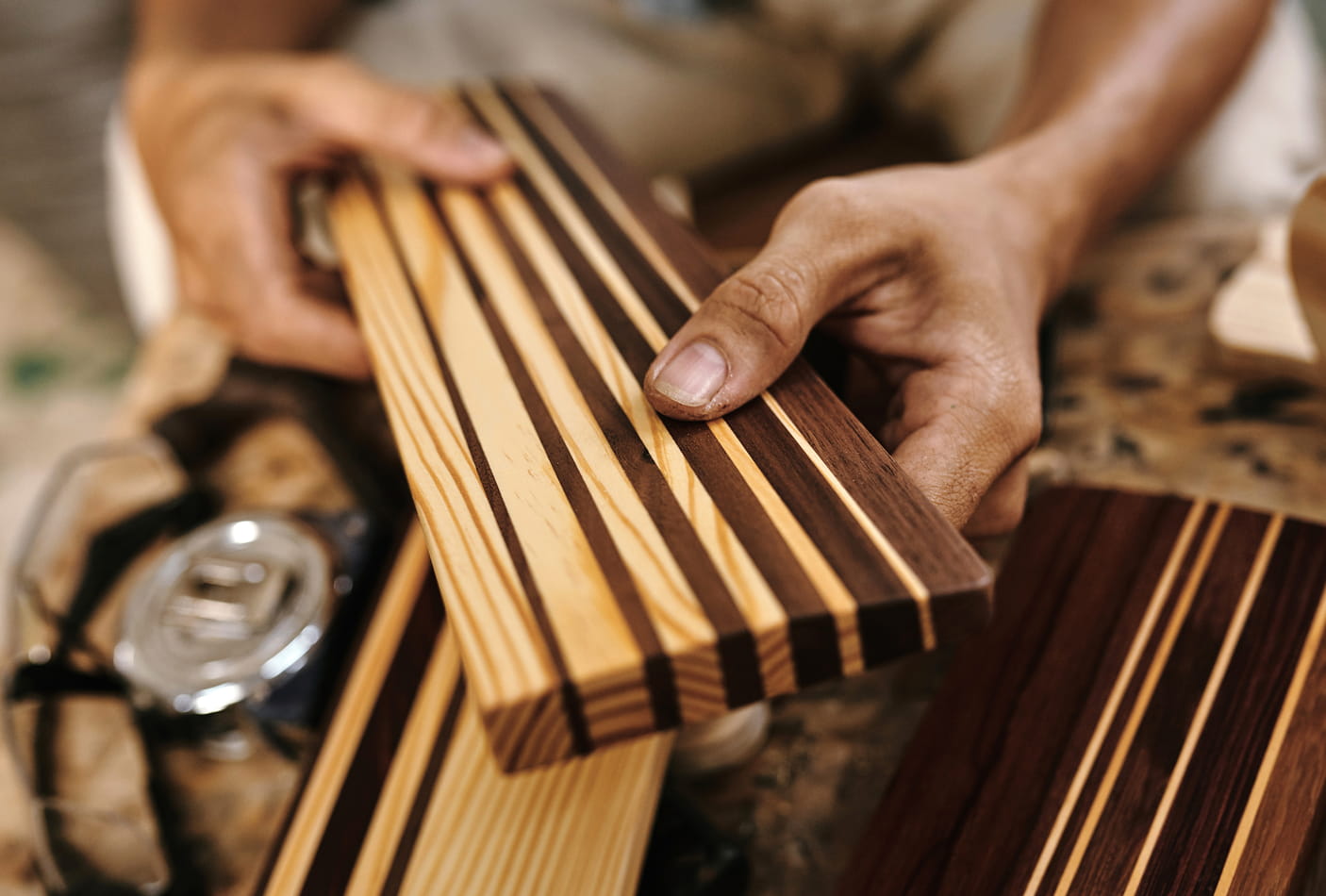 Close-up of hands holding a piece of polished, striped wood with various shades of brown and beige in a woodworking workshop.