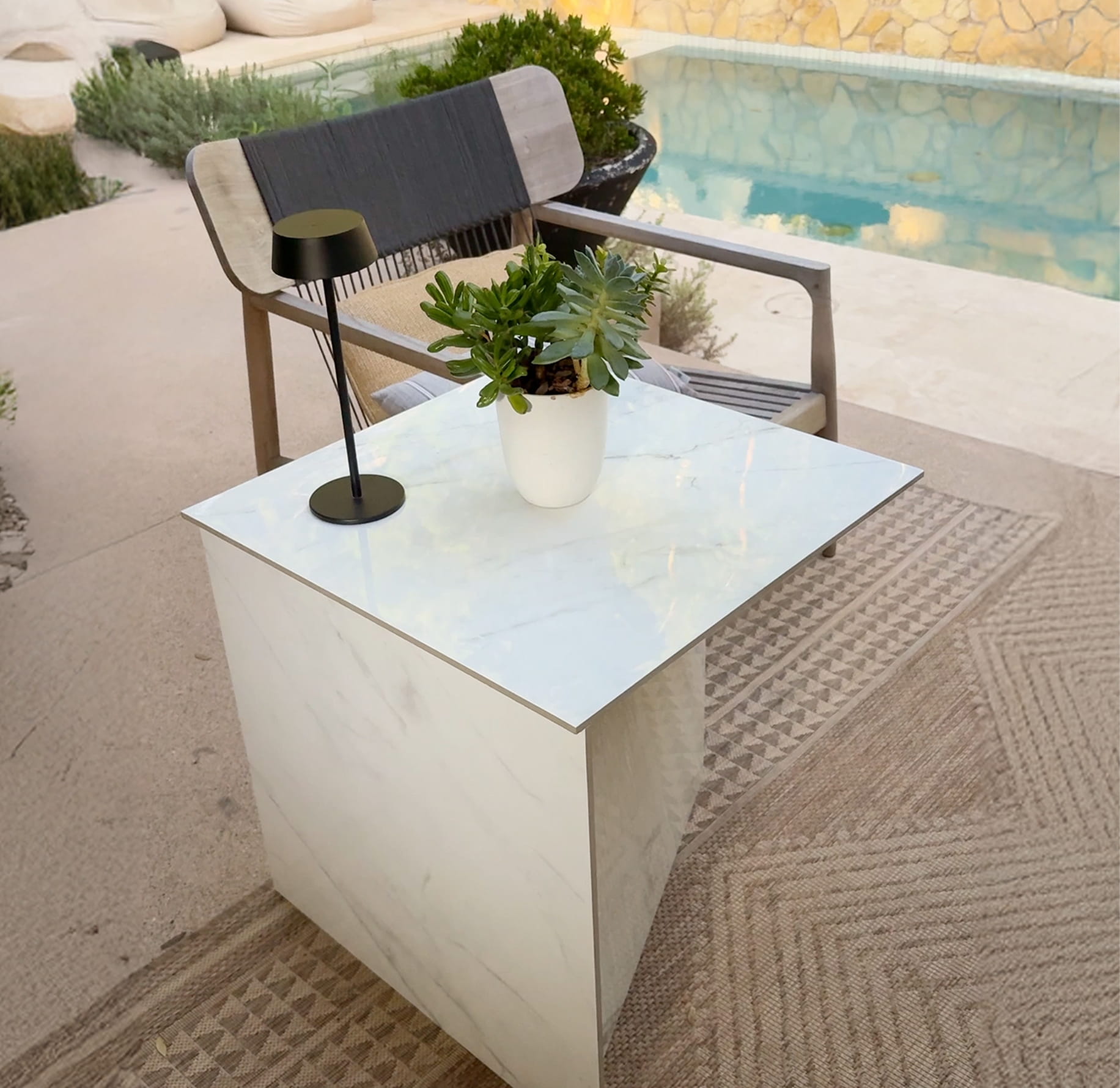 White square marble side table with a black modern lamp and a white pot with succulents next to a wooden chair by a pool.