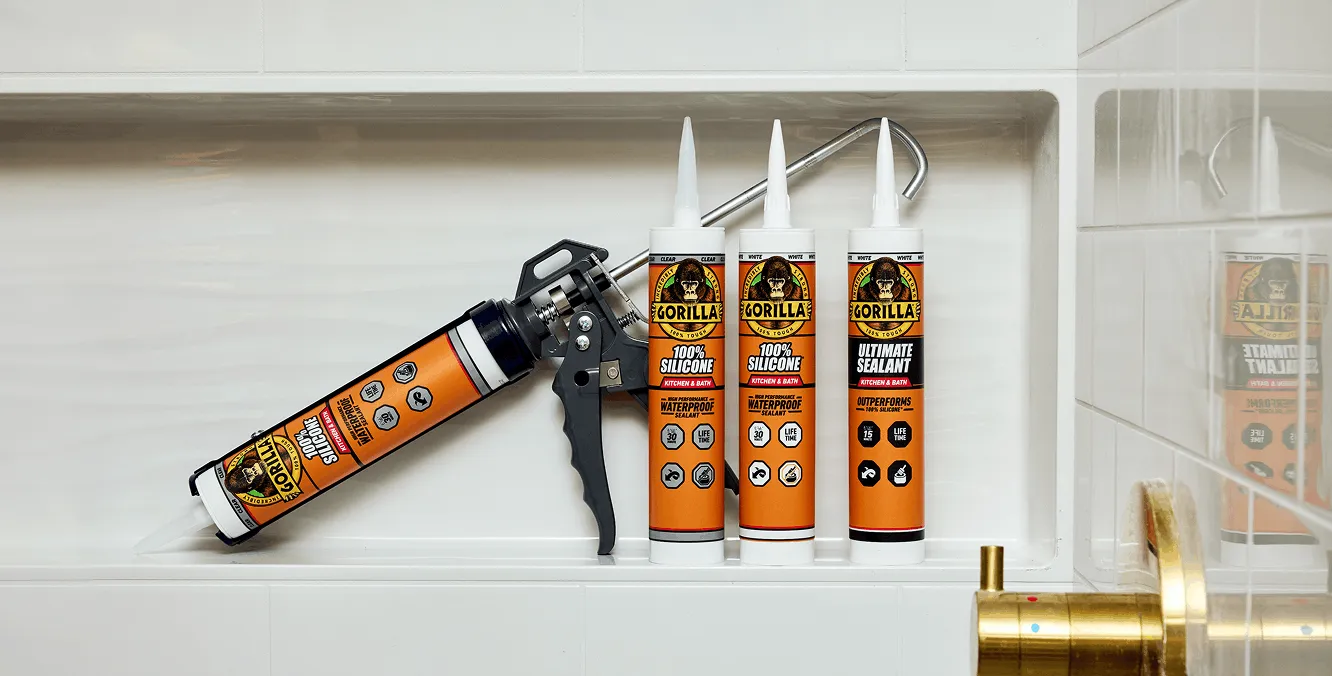 Four tubes of Gorilla silicone sealant with a caulking gun on a white tiled shower shelf near a brass faucet.