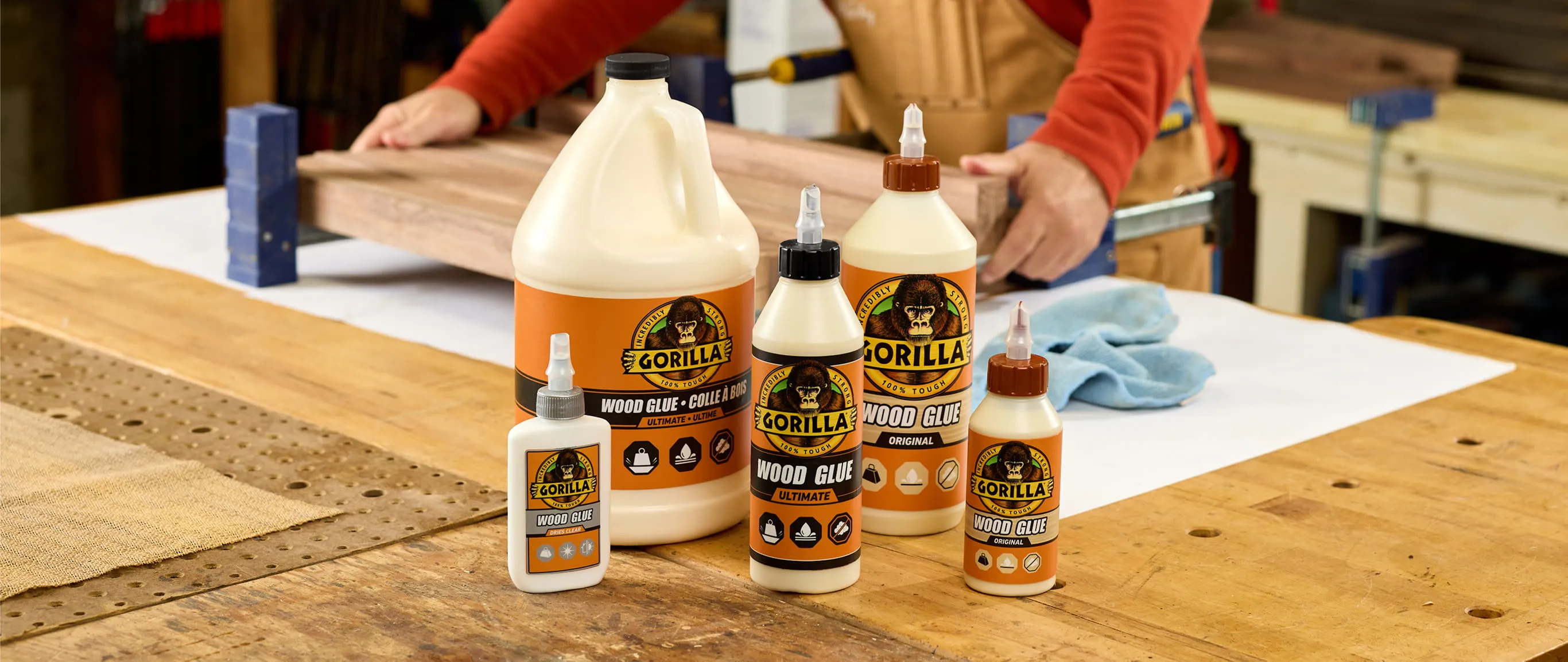 Various sizes of Gorilla wood glue bottles on a woodworking bench with a person working on wood in the background.