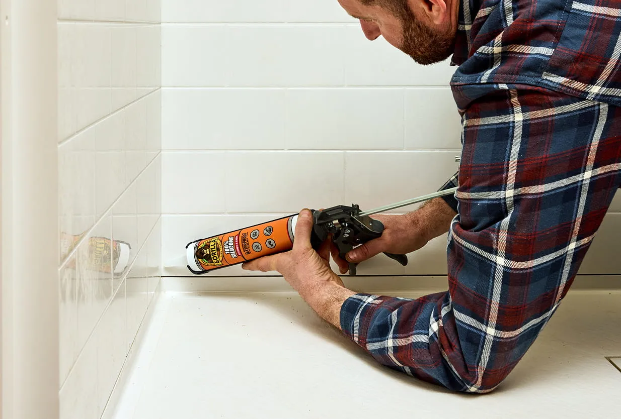 Man in plaid shirt applying Gorilla waterproof silicone sealant with a caulking gun in a shower.