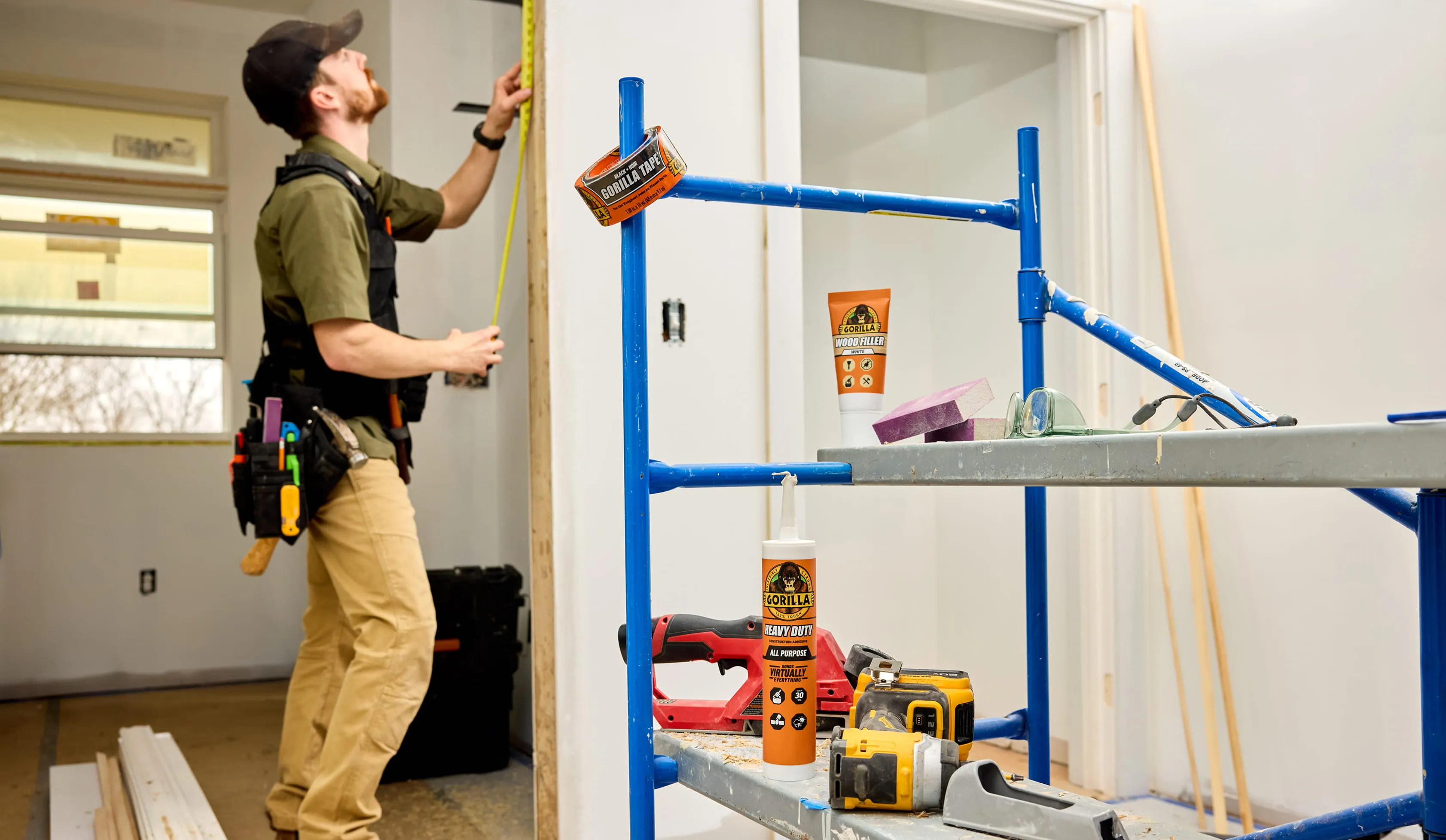 Contractor measuring a wall with a tape measure near scaffolding with Gorilla brand construction adhesive, wood filler, and tools in the foreground.