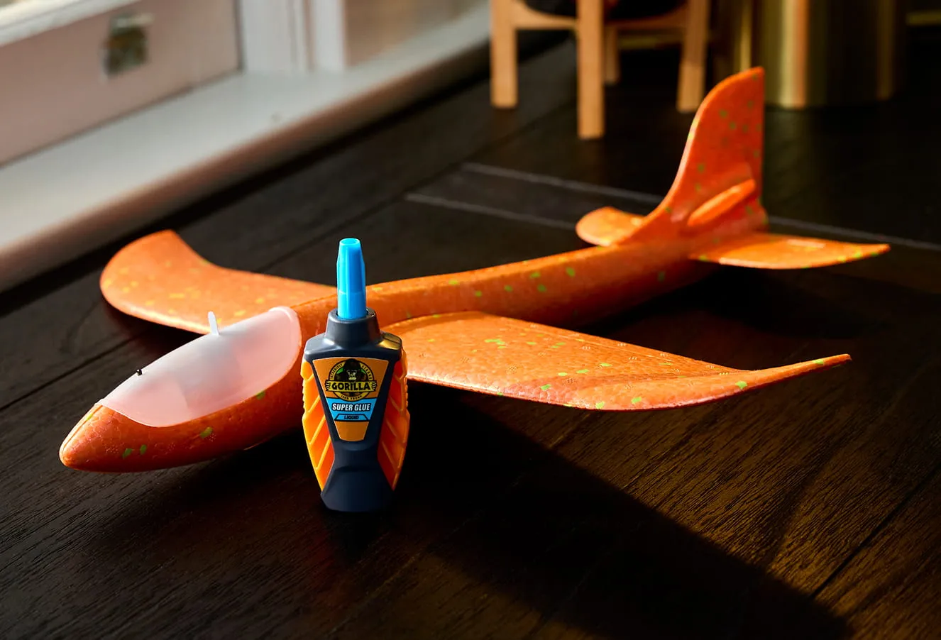 Bottle of Gorilla Super Glue placed in front of an orange foam model airplane on a dark wooden surface.
