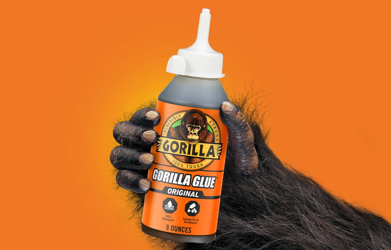 Gorilla hand holding an 8-ounce bottle of Gorilla Glue Original against an orange background.