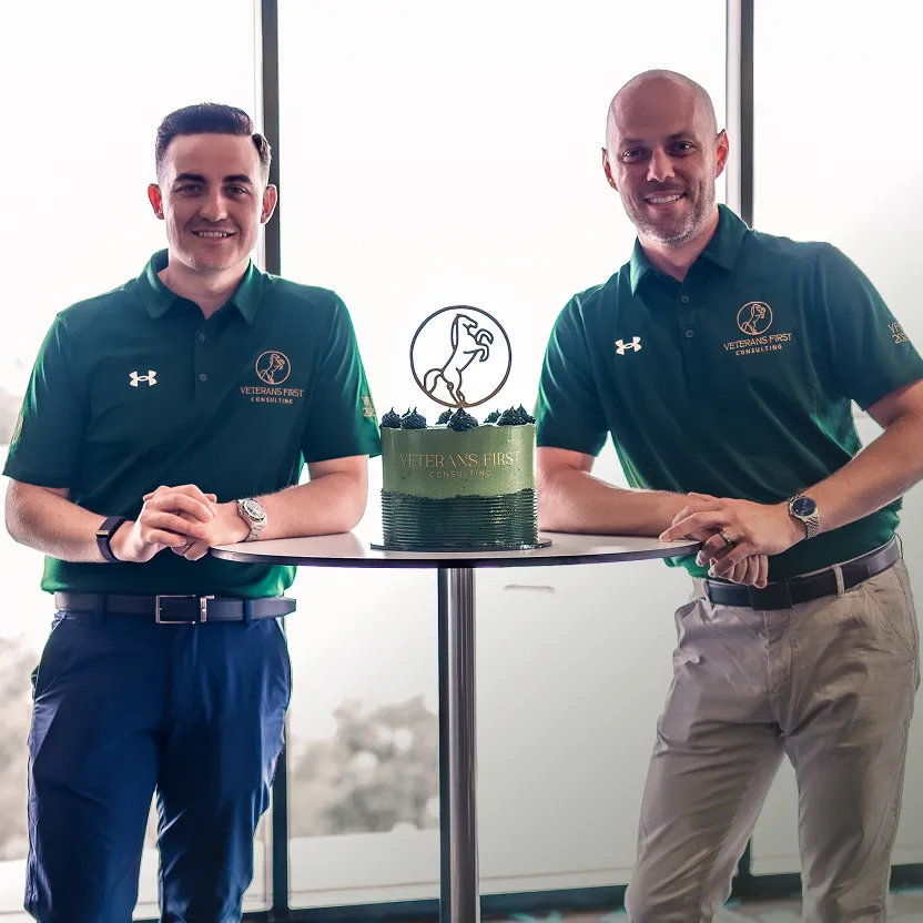 Two men wearing green Veterans First Consulting polo shirts standing on either side of a cake on a round table.