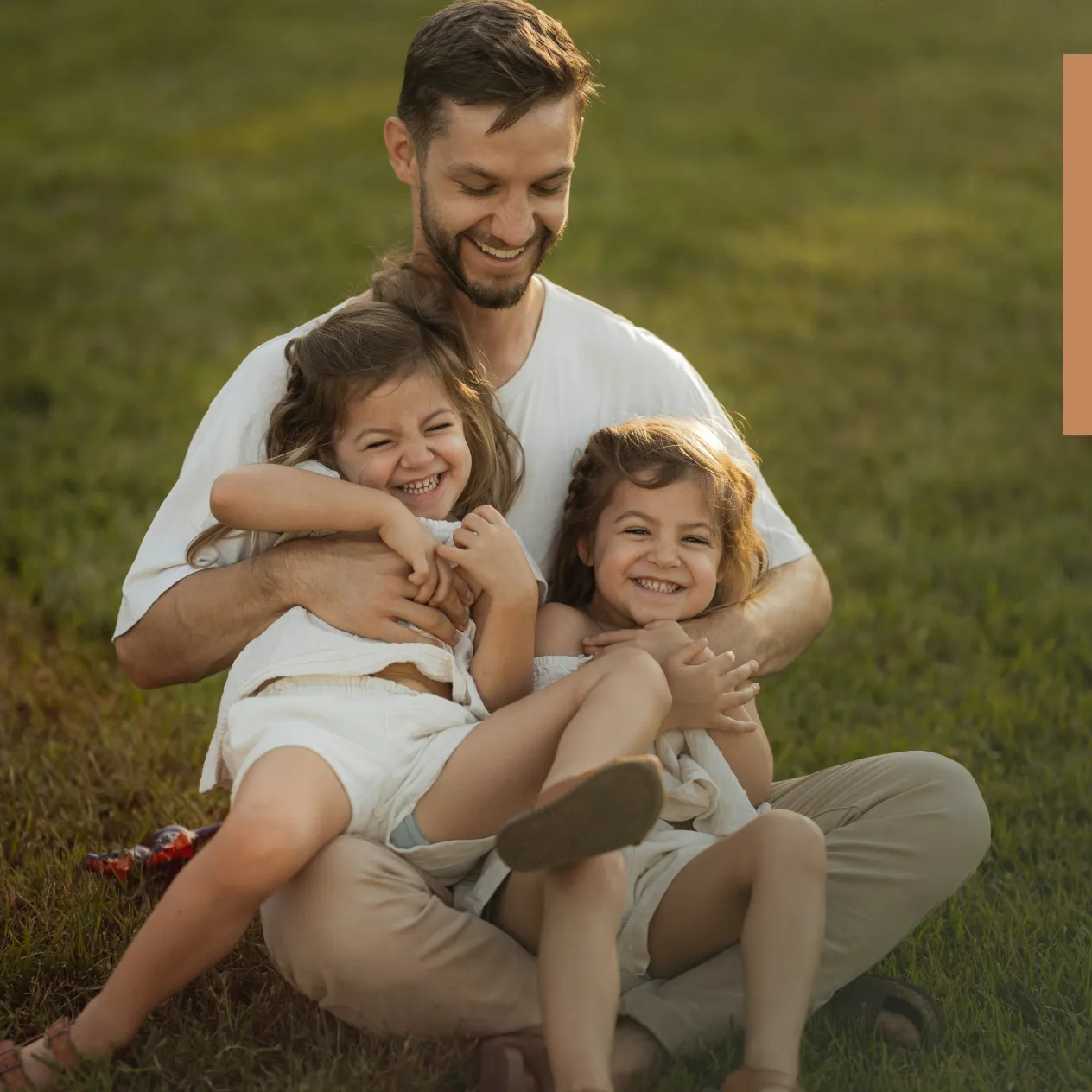 Smiling man sitting on grass outdoors, hugging and tickling two laughing young girls.