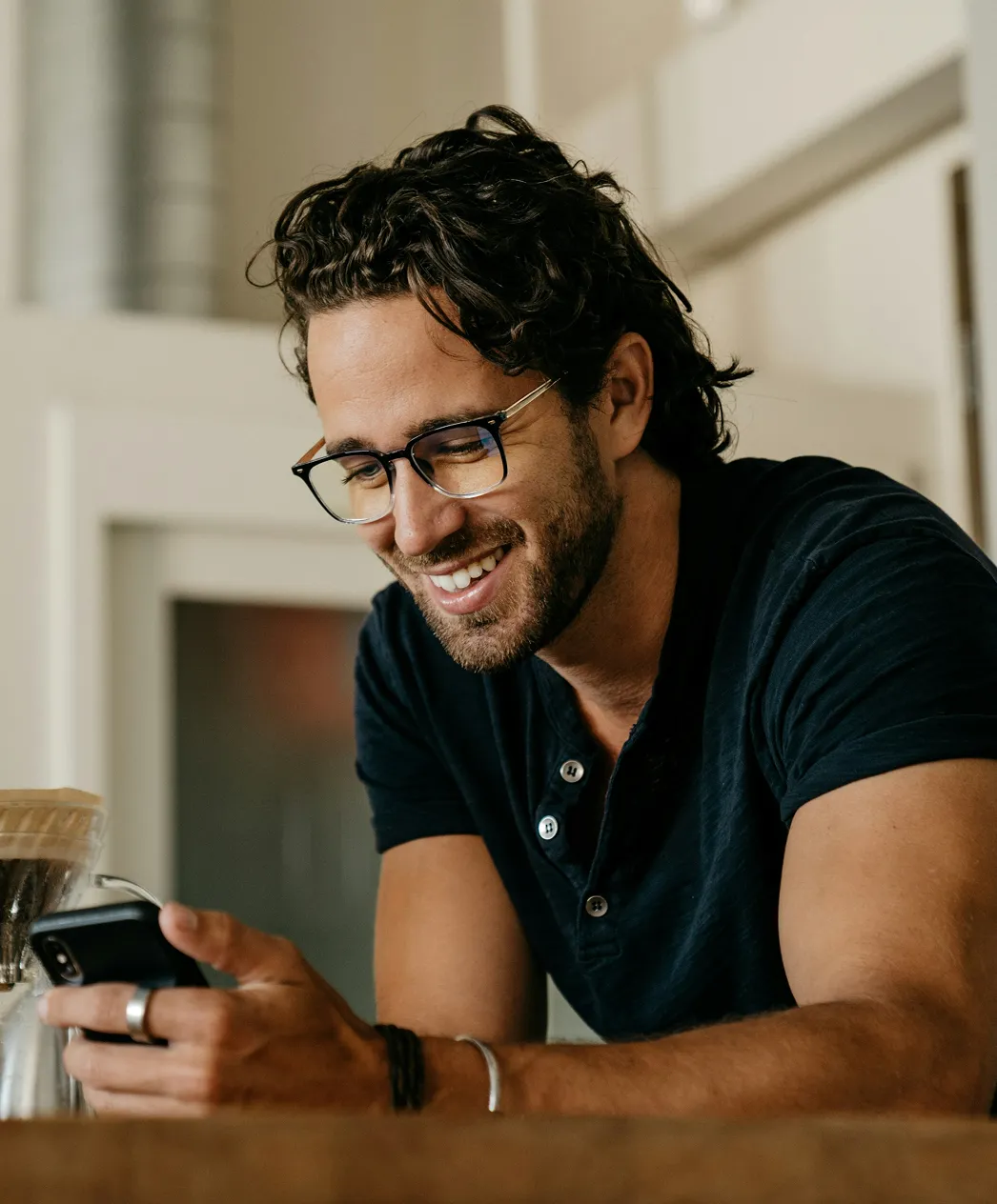 Smiling man with curly hair and glasses looking at his smartphone indoors.