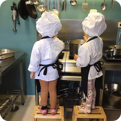 Barn klädda i kockkläder lagar mat tillsammans i köket, en lekfull aktivitet som främjar kreativitet och samarbete.