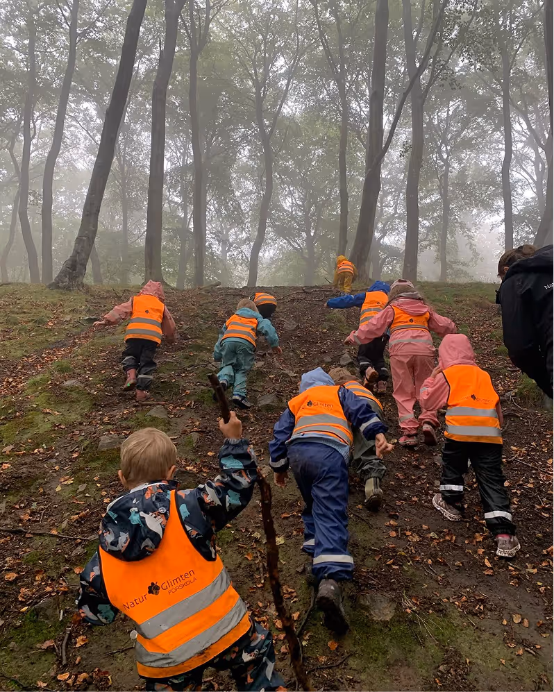 Barn i färgglada kläder och orange reflexvästar klättrar uppför en lerig kulle i en tät dimmig skog.