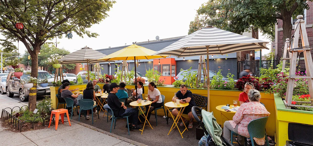 Outdoor café scene with patrons seated at colorful tables under umbrellas, surrounded by greenery and urban settings.