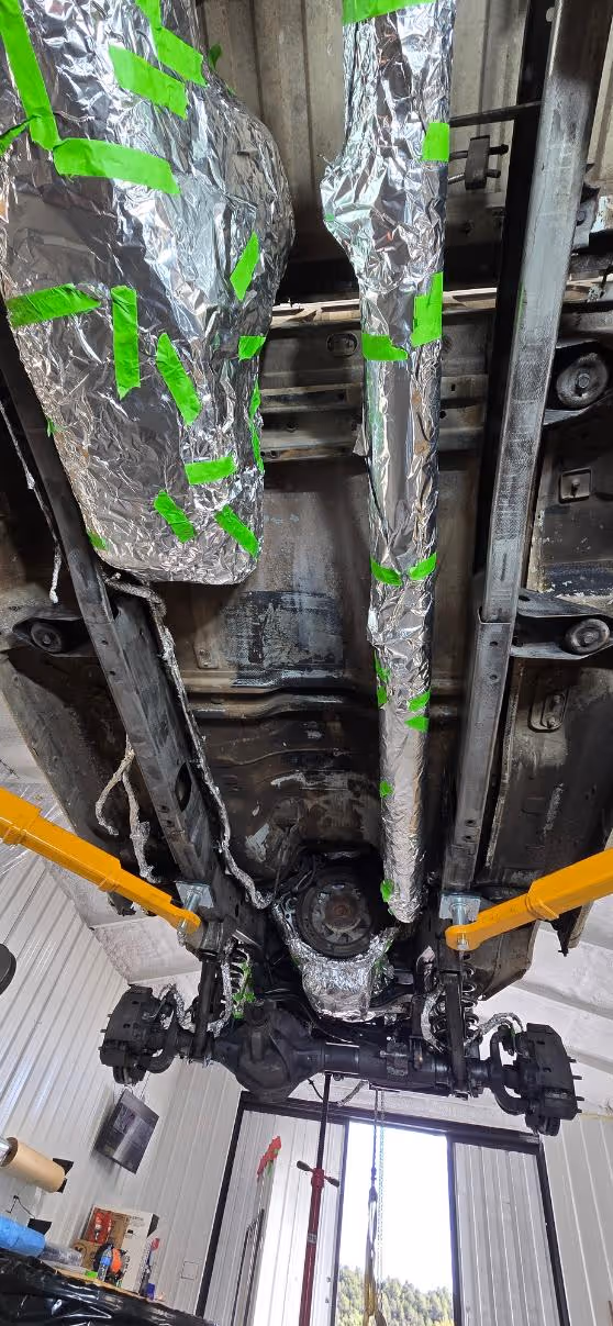 Underside of a vehicle on a lift in a garage with parts wrapped in foil and green tape for rust removal preparation.