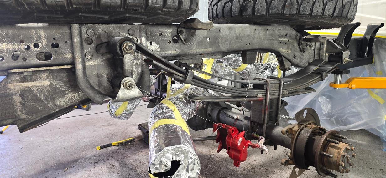 Underside view of a truck bed chassis undergoing rust removal with wrapped exhaust pipes and a red brake caliper.