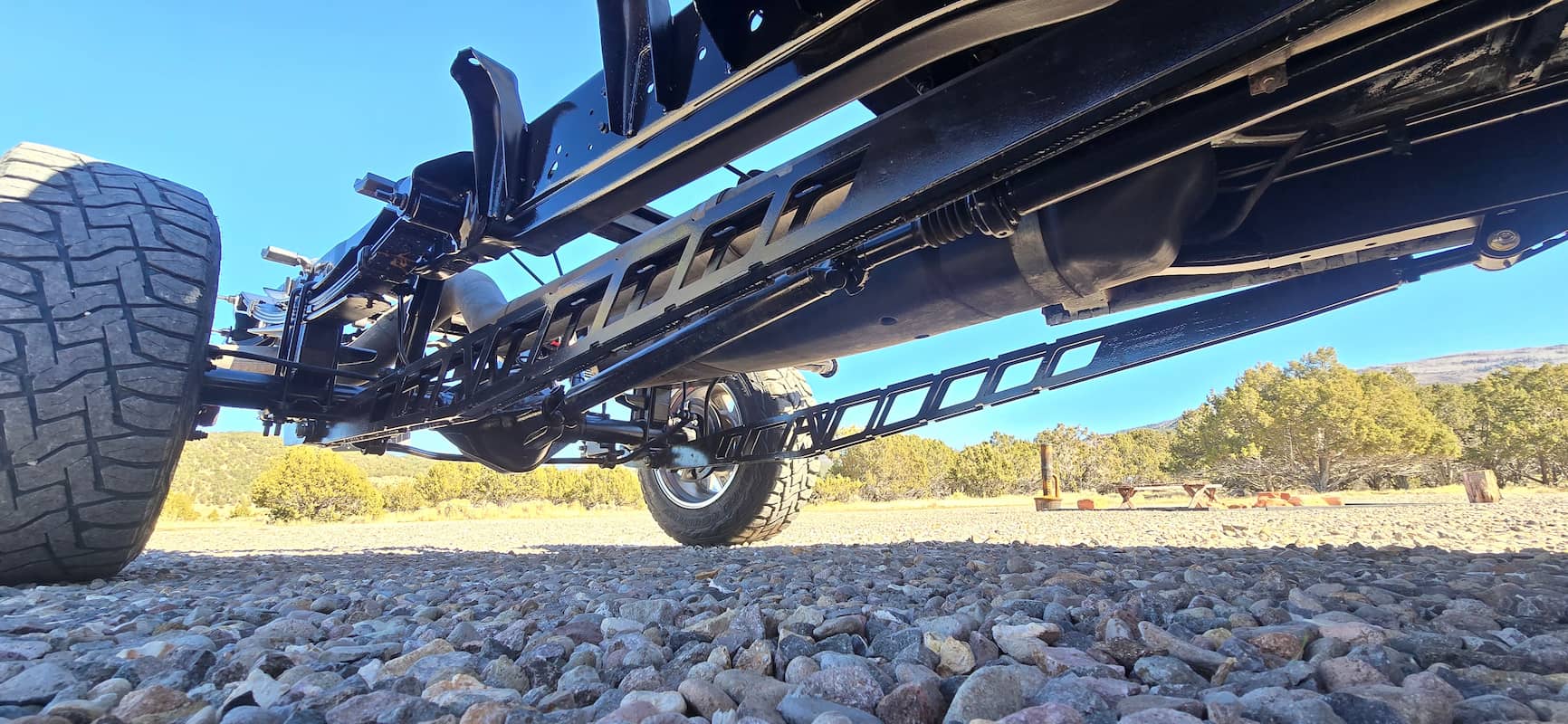 Underside view of a black truck chassis with large off-road tires on rocky ground, trees and clear sky in background.
